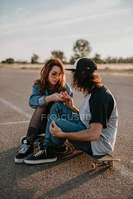 Модная подростковая пара, сидящая на коньках на пустой дороге и разговаривающая, глядя друг на друга — стоковое фото
