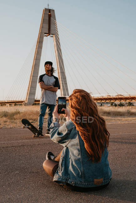 Long haired stylish teenage female shooting cool hipster guy with skateboard against modern bridge in countryside — Stock Photo