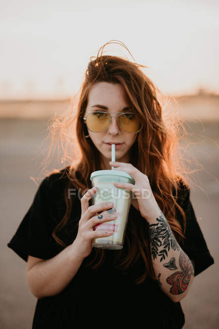 Очаровательная модная девушка-подросток в солнечных очках и с татуировками, наслаждающаяся молочным коктейлем из стекла с соломинкой, улыбающейся в камеру на закате — стоковое фото