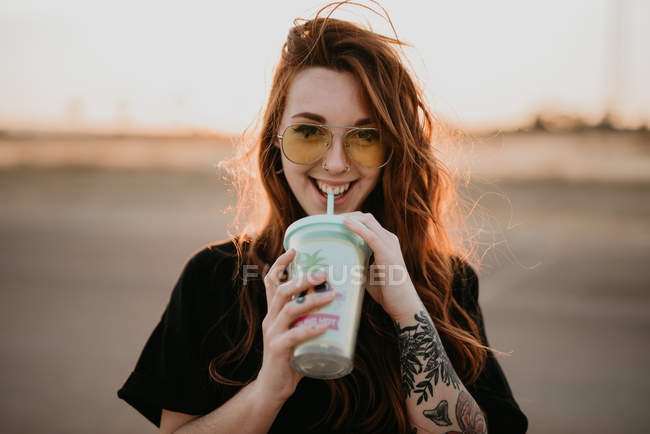 Charmante trendige Teenager-Mädchen mit Sonnenbrille und Tätowierungen genießen Milchshake aus Glas mit Stroh lächelnd in die Kamera im Sonnenuntergang — Stockfoto