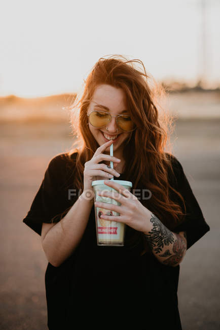 Очаровательная модная девушка-подросток в солнечных очках и с татуировками, наслаждающаяся молочным коктейлем из стекла с соломой на закате — стоковое фото