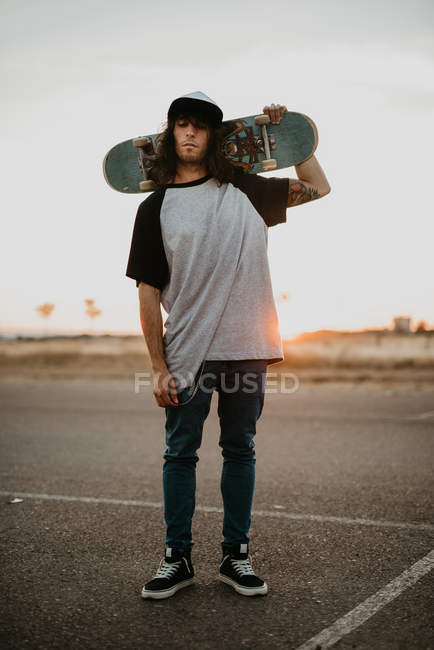 Стильный хипстер-подросток, держащий скейтборд на затылке и смотрящий в камеру на пустой дороге на закате — стоковое фото