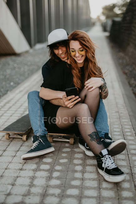 Крута модна пара сидить на дорозі зі скейтбордом, використовуючи мобільний телефон у місті — стокове фото