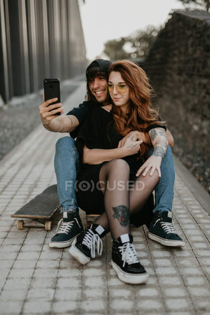 Cool trendy couple sitting on road with skateboard and taking selfie together with mobile phone in city — Stock Photo