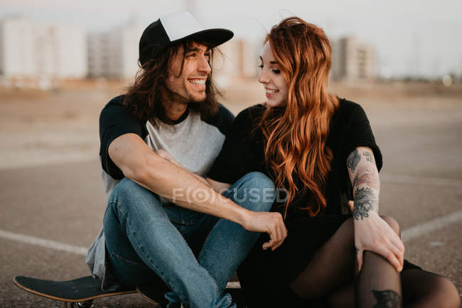 Trendy adolescente coppia seduta su pattini sulla strada remota vuota e parlando guardarsi l'un l'altro — Foto stock