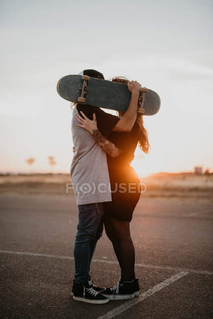 Side view of romantic teenage couple embracing and covering heads with skateboard while kissing in back lit of sunset on rural road — Stock Photo