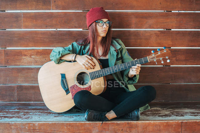 Стильный подросток задумчиво играет на гитаре, сидя на земле со скрещенными ногами и отводя взгляд — стоковое фото