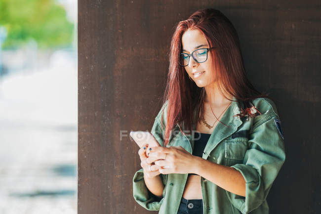 Long haired teenager in glasses surfing mobile phone nearby wall in bright day — Stock Photo