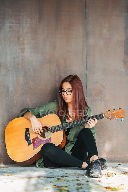 Стильный подросток задумчиво играет на гитаре, сидя на земле со скрещенными ногами — стоковое фото