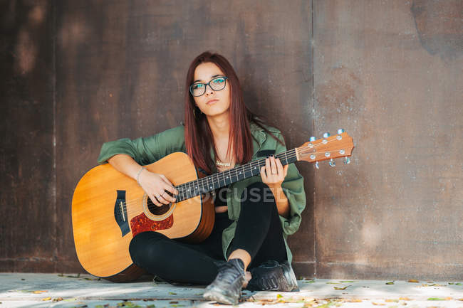 Стильний зміст підліток задумливо грає на гітарі, сидячи на землі з схрещеними ногами і дивлячись на камеру — стокове фото