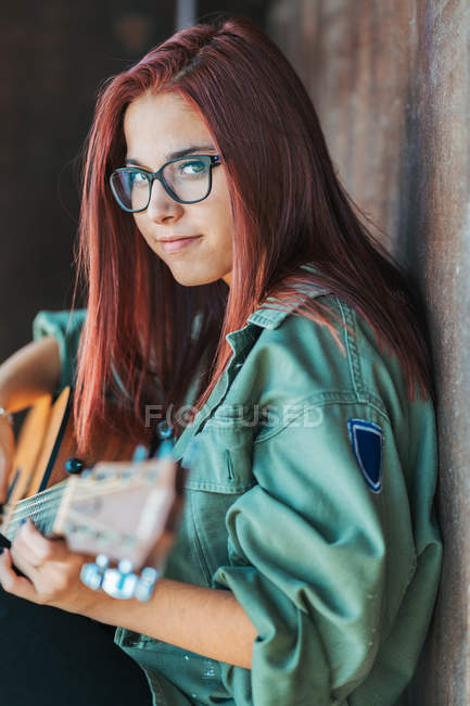 Vista lateral de conteúdo elegante adolescente tocando violão sentado no chão com pernas cruzadas e olhando para a câmera — Fotografia de Stock