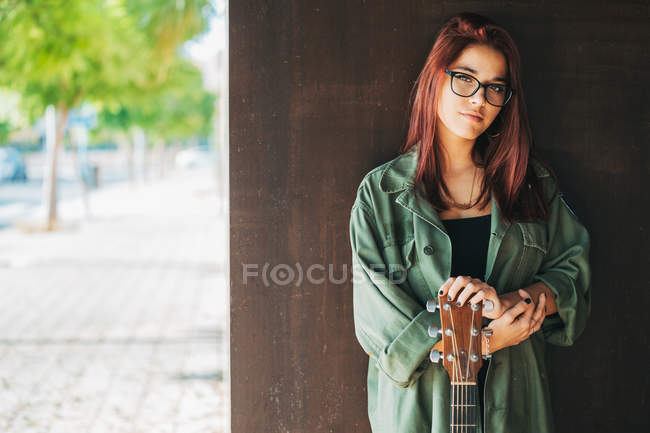Content stylish teenager in glasses in dark green shirt holding a guitar standing nearby brown wall looking at camera — Stock Photo