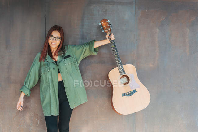 Содержание стильный подросток в очках в темно-зеленой рубашке держа гитару, стоящую рядом коричневая стена, глядя на камеру — стоковое фото