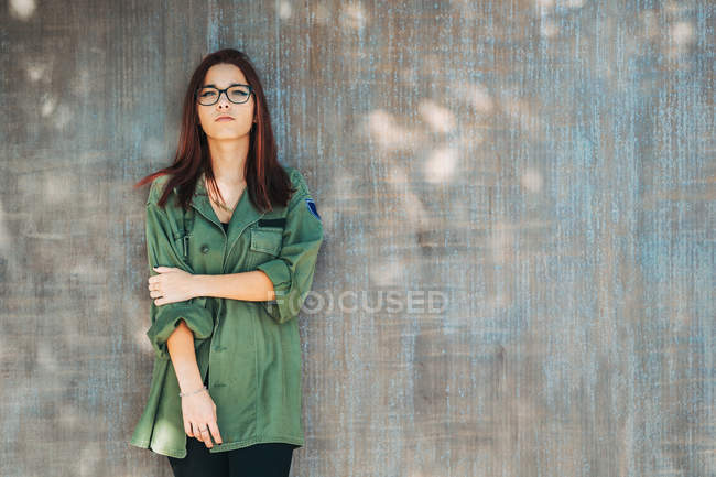 Contenido adolescente con estilo en gafas en camisa verde oscuro cerca de la pared marrón mirando a la cámara - foto de stock