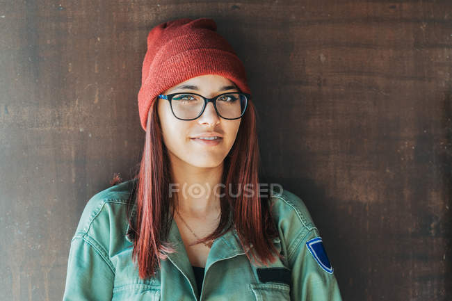 Nachdenklich stilvoller Teenager mit warmem Hut und Brille in dunkelgrünem Hemd neben brauner Wand, der mitschaut — Stockfoto