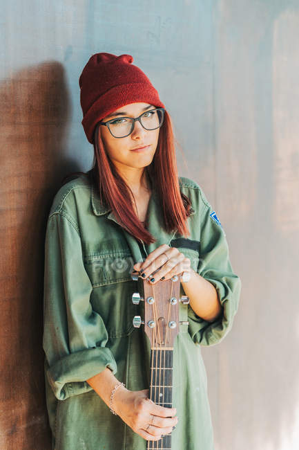 Content stylish teenager in glasses in dark green shirt holding a guitar standing nearby brown wall looking at camera — Stock Photo