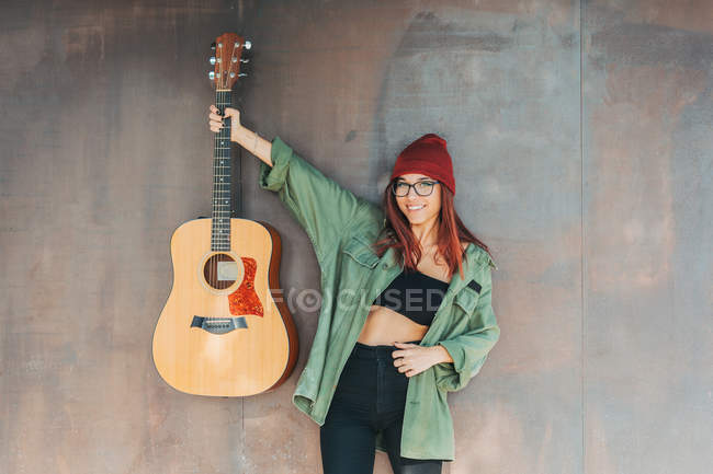 Contenido adolescente con estilo en gafas en camisa verde oscuro sosteniendo una guitarra de pie cerca de la pared marrón mirando a la cámara - foto de stock