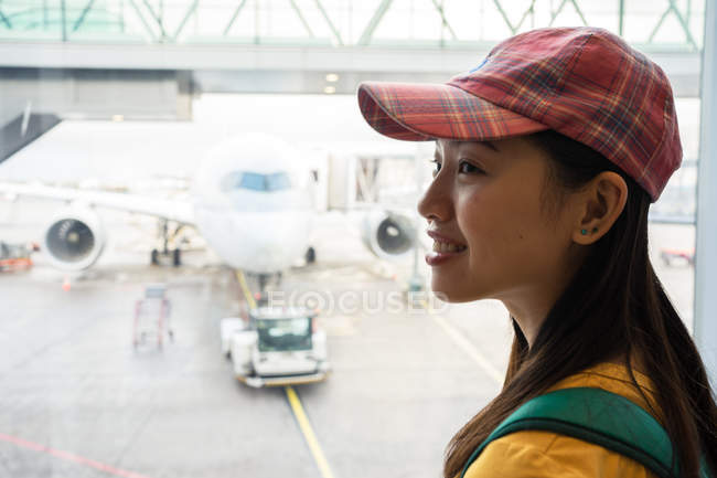 Вид сбоку жизнерадостной азиатской женщины в кепке у окна с видом на взлетно-посадочную полосу с самолетом и автопогрузчиком — стоковое фото