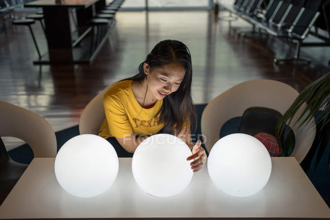Langhaarige aufgeregte asiatische Frau beobachtet und berührt weiße leuchtende runde Lampen auf dem Tisch in der Wartehalle — Stockfoto