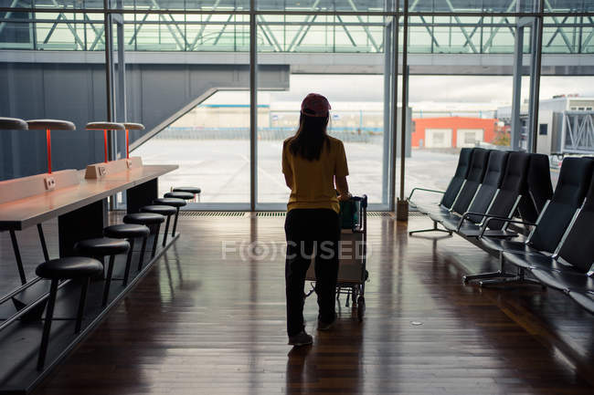 Visão traseira da mulher em boné e desgaste esportivo andando com bagagem ao longo de cadeiras no aeroporto — Fotografia de Stock