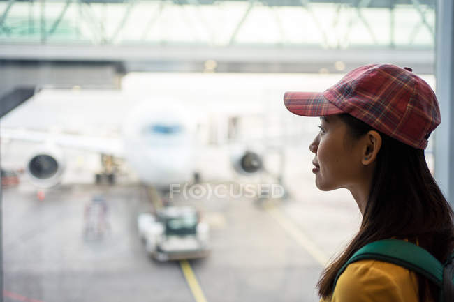 Сторона зору азіатської жінки в шапці на вікні з видом на злітно-посадкову смугу з літаком і завантажувачем автомобілів. — стокове фото