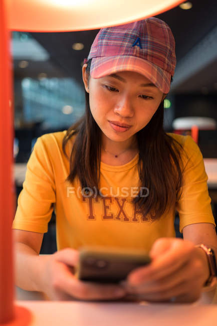 Mujer asiática en gorra surfeando en el teléfono móvil sentado en la mesa antes de la tabla de horarios en el aeropuerto - foto de stock