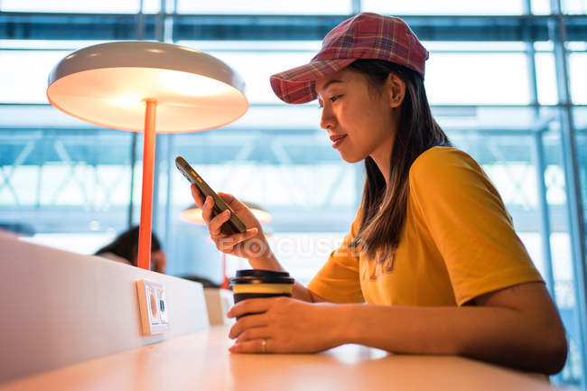 Side view of asian woman in cap surfing mobile phone and drinking coffee from disposable cap at table in airport — Stock Photo