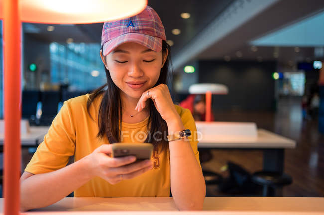 Asian woman in cap surfing on mobile phone sitting at table before schedule board in airport — Stock Photo