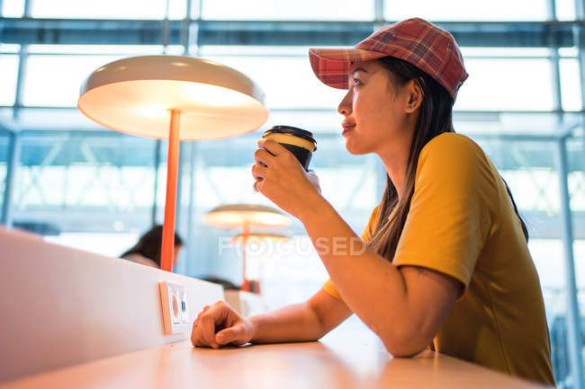 Vista lateral da mulher asiática em cap beber café de tampa descartável na mesa com iluminação e olhando para cima no aeroporto — Fotografia de Stock