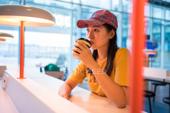Side view of Asian woman in cap drinking coffee from disposable cap at table with illumination and looking up in airport — Stock Photo