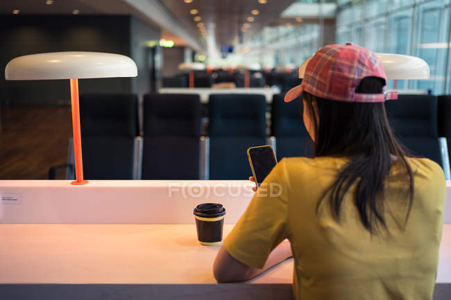Rückansicht einer Frau mit Schirmmütze, die am Tisch im Flughafen Handy surft und Kaffee aus einer Einwegkappe trinkt — Stockfoto