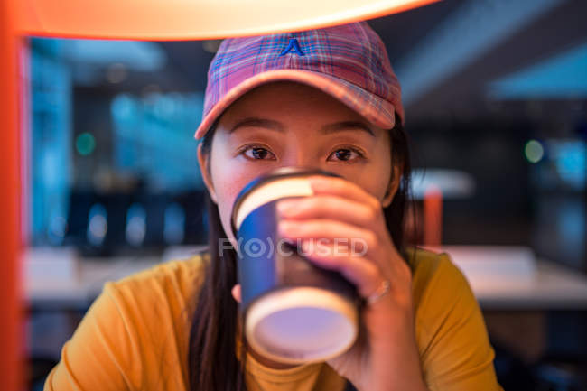Asiatin in Mütze trinkt Kaffee aus Einwegdeckel am Tisch mit Beleuchtung im Flughafen — Stockfoto