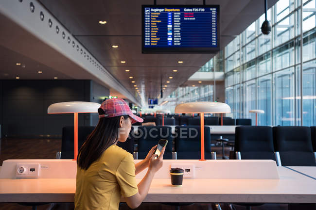 Vista lateral da mulher asiática em cap surf telefone celular e beber café de tampa descartável na mesa no aeroporto — Fotografia de Stock