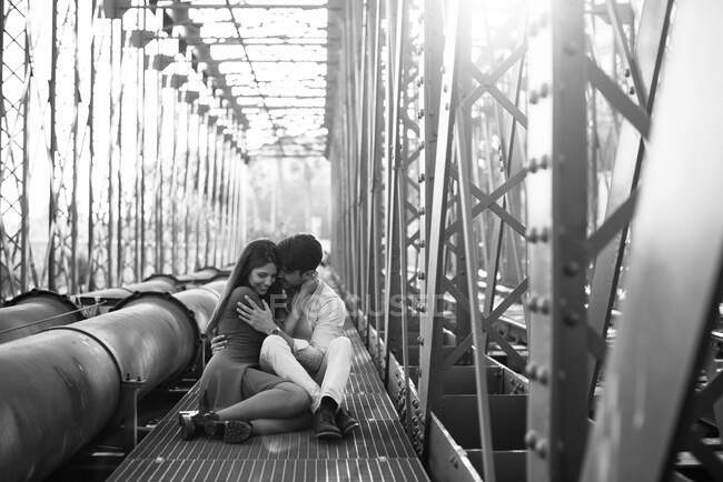 Hombre cariñoso y mujer tierna en vestido azul abrazándose en el ferrocarril bajo construcción de puente de metal - foto de stock