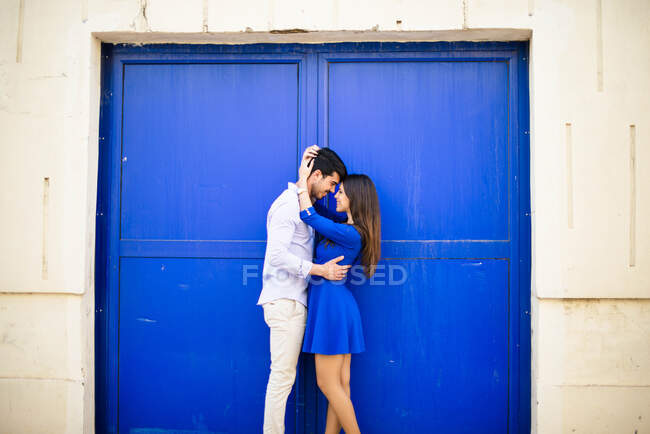 Vue latérale de l'homme et de la femme aimants dans des vêtements élégants embrasser sur fond de porte bleu vif — Photo de stock