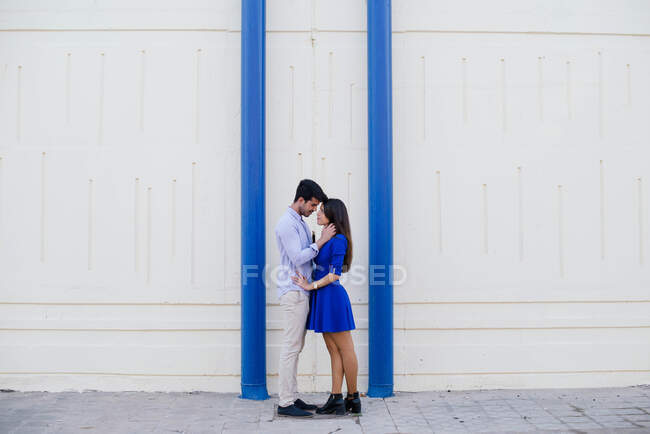 Vista lateral de hombre y mujer cariñosos en ropa elegante besándose en el fondo de la pared blanca con columnas azules brillantes - foto de stock