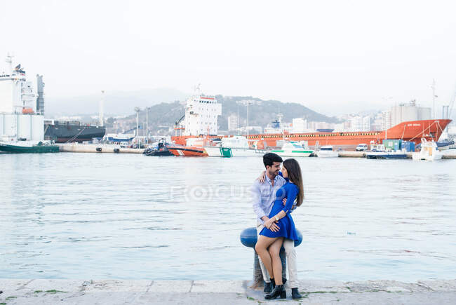 Вид сбоку на очаровательную женщину в синем платье в объятиях красивого мужчины, скрепляющегося на пирсе городского порта на фоне корабля — стоковое фото