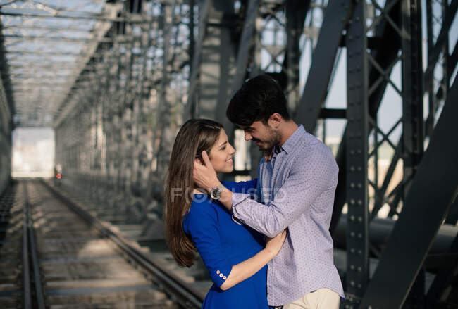 Люблячий чоловік і ніжний жінка в блакитній сукні, обіймаючись залізницею під будівництвом металевого мосту — стокове фото