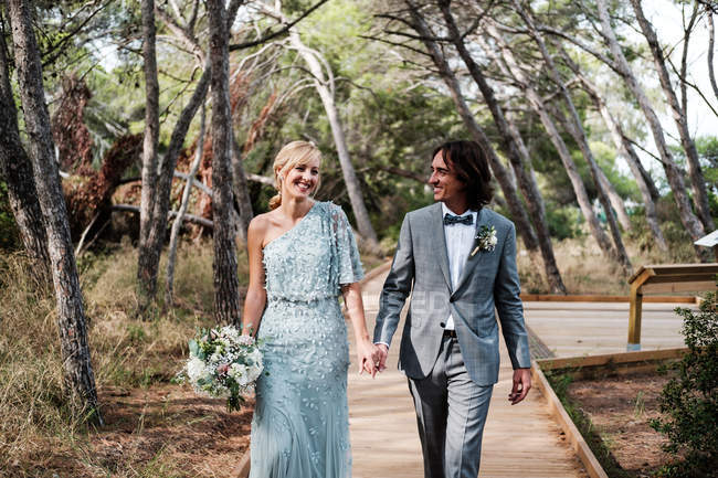 Jovem casal recém-casado em casamento desgaste andando, de mãos dadas no caminho entre bela floresta verde com árvores grandes — Fotografia de Stock