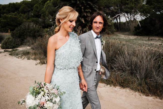 Beautiful newly married couple in stylish wedding clothing holding hands while walking on nature — Stock Photo