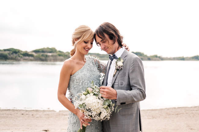 Freut sich junger Bräutigam im Hochzeitsanzug, der stolz auf die schöne blondhaarige Braut im stylischen Kleid hinter dem Ufer blickt — Stockfoto
