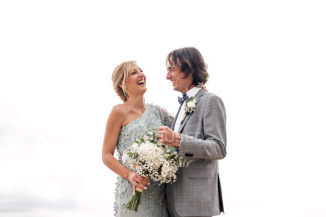 Задоволений молодий наречений у весільному костюмі гордо дивиться на красиву блондинку з волоссям наречену в стильній сукні позаду — стокове фото