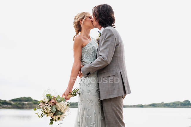 Pleased young groom in wedding suit hugging and kissing blonde haired bride in stylish dress behind at seashore — Stock Photo