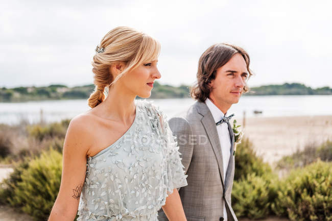 Young pleased happy couple in wedding day walking throughout empty sandy seashore — Stock Photo