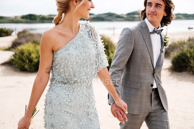 Задоволений молодий наречений у весільному костюмі гордо дивиться на красиву блондинку з волоссям наречену в стильній сукні позаду на березі моря — стокове фото