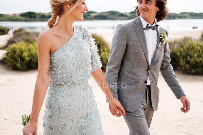 Agradable novio joven en traje de novia con orgullo mirando hermosa novia de pelo rubio en vestido elegante detrás en la orilla del mar - foto de stock