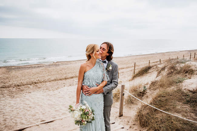Zufriedener junger Bräutigam im Hochzeitsanzug umarmt und küsst blondhaarige Braut im stilvollen Kleid hinter leeren Sandstränden — Stockfoto