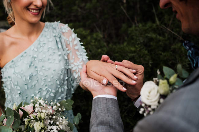 Cropped image of bridegroom in wedding suit putting ring on finger of happy future wife with green plants on background — Stock Photo