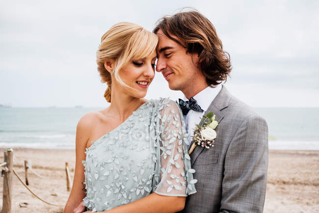 Zufriedener junger Bräutigam im Hochzeitsanzug umarmt blondhaarige Braut im stylischen Kleid hinter leeren Sandstränden — Stockfoto