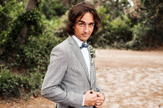 Young groom in stylish gray wedding suit going on empty road with green trees on sides — Stock Photo